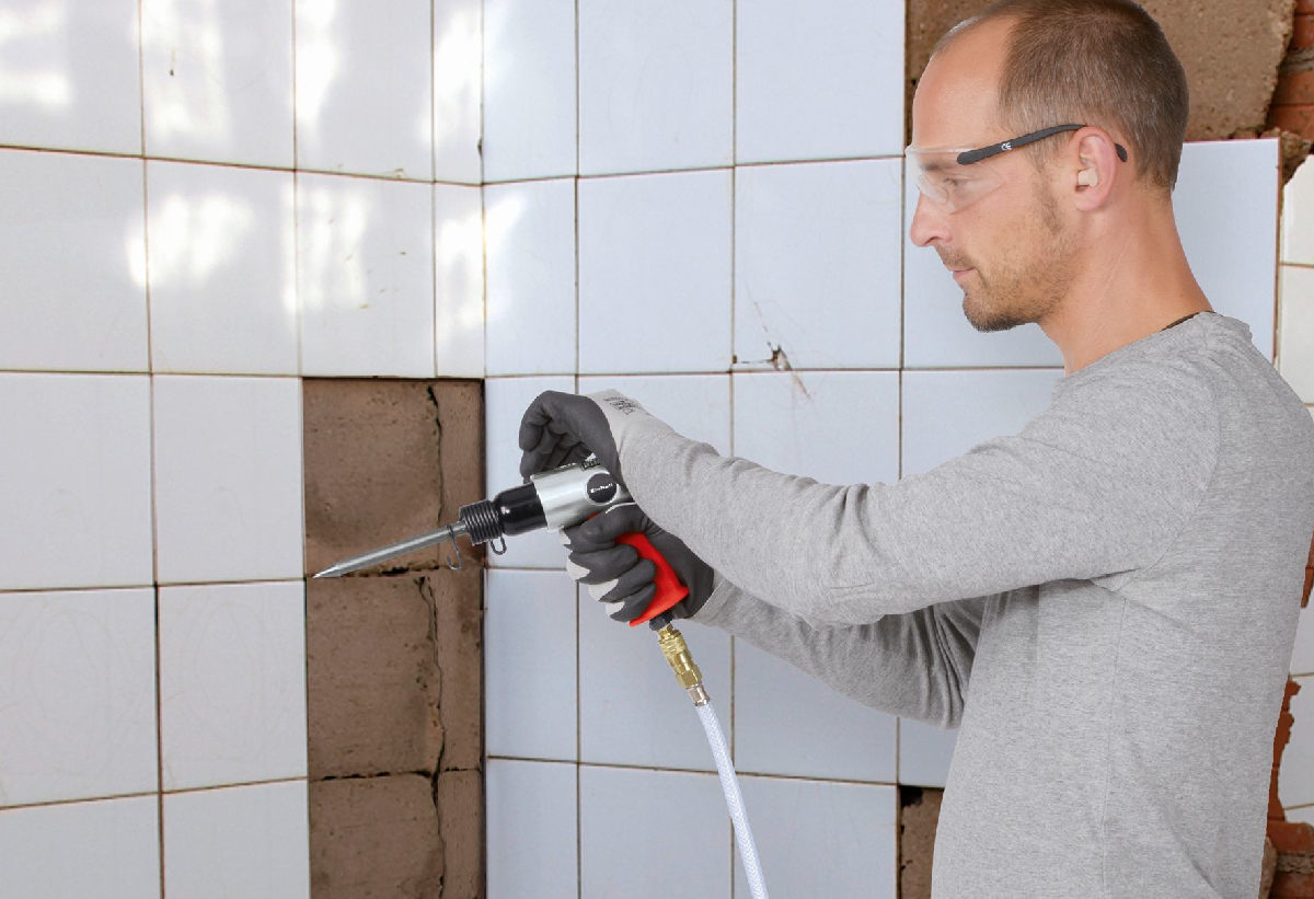 Απεικονίζεται άνδρας να χρησιμοποιεί τον αεροσυμπιετή σε έναν τοίχο με λευκά πλακάκια.